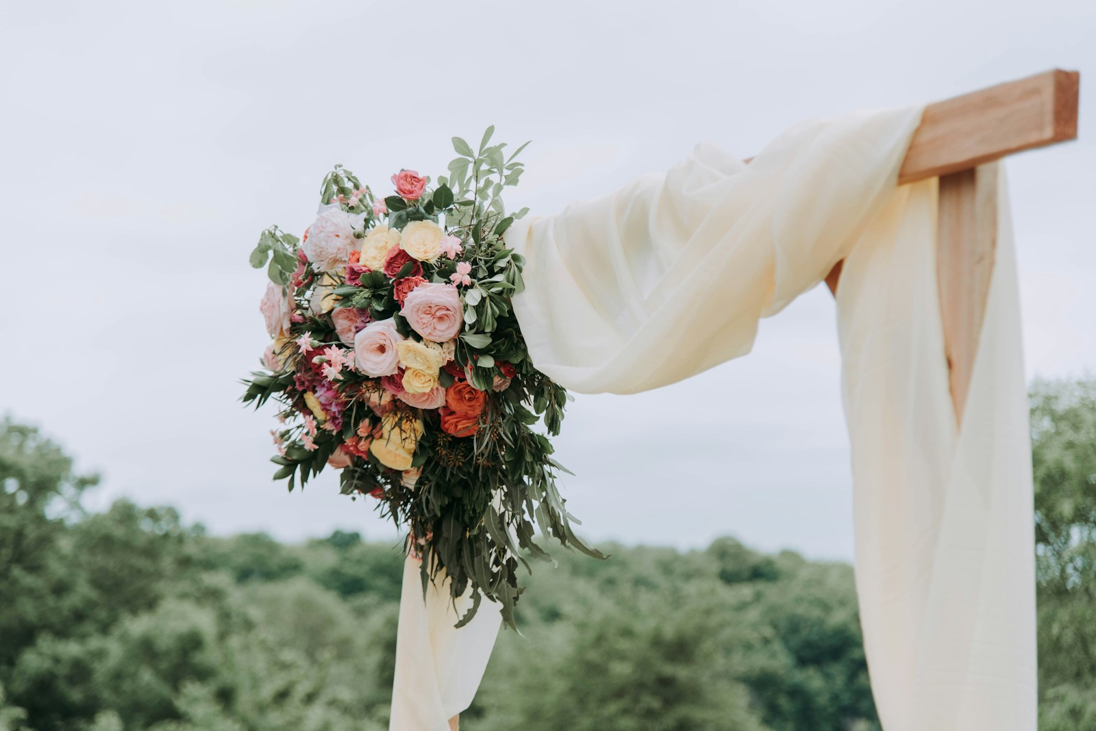 A chuppah corner: a wooden post with draped cream fabric and flowers, trees behind